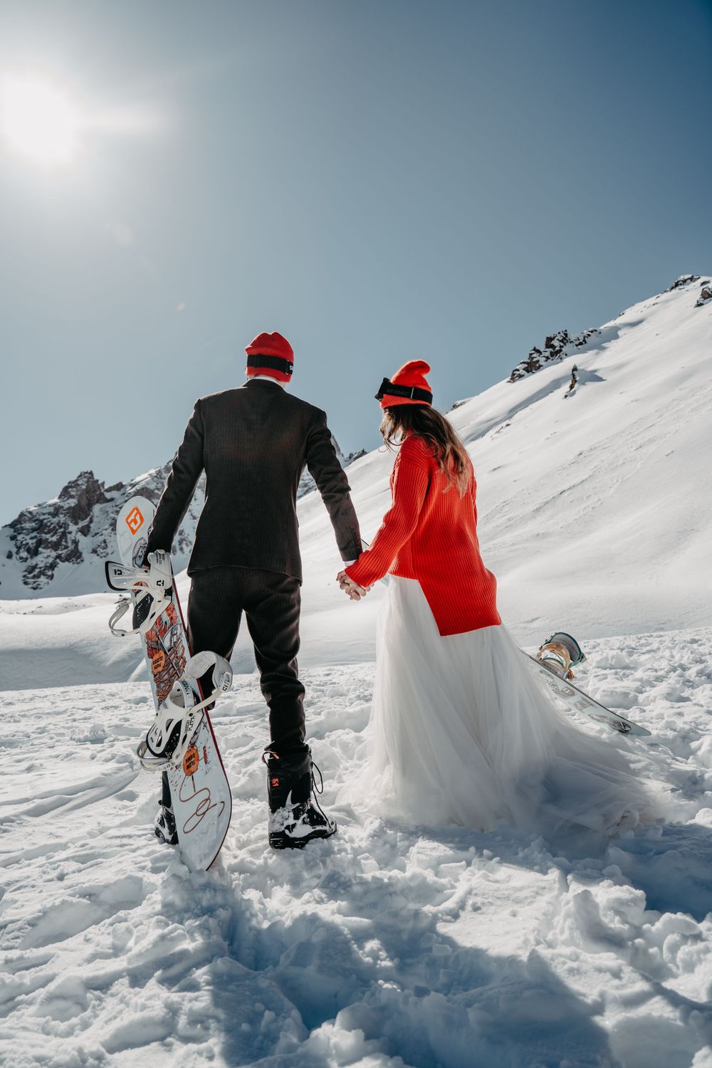 Bride and groom walking in snowy mountains carrying a snowboard