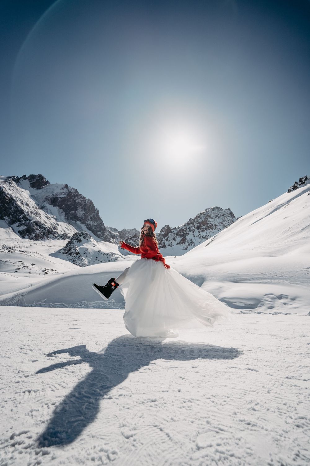 A girl wearing a red sweater over a wedding dress walking in snowy mountains