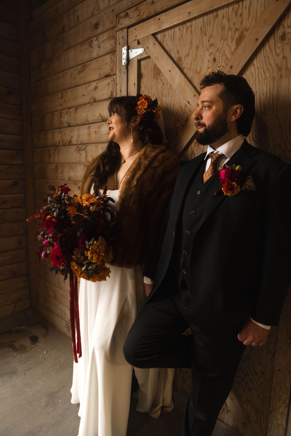 A couple in formal wedding attire stands intimately together against a rustic wooden wall in moody lighting.