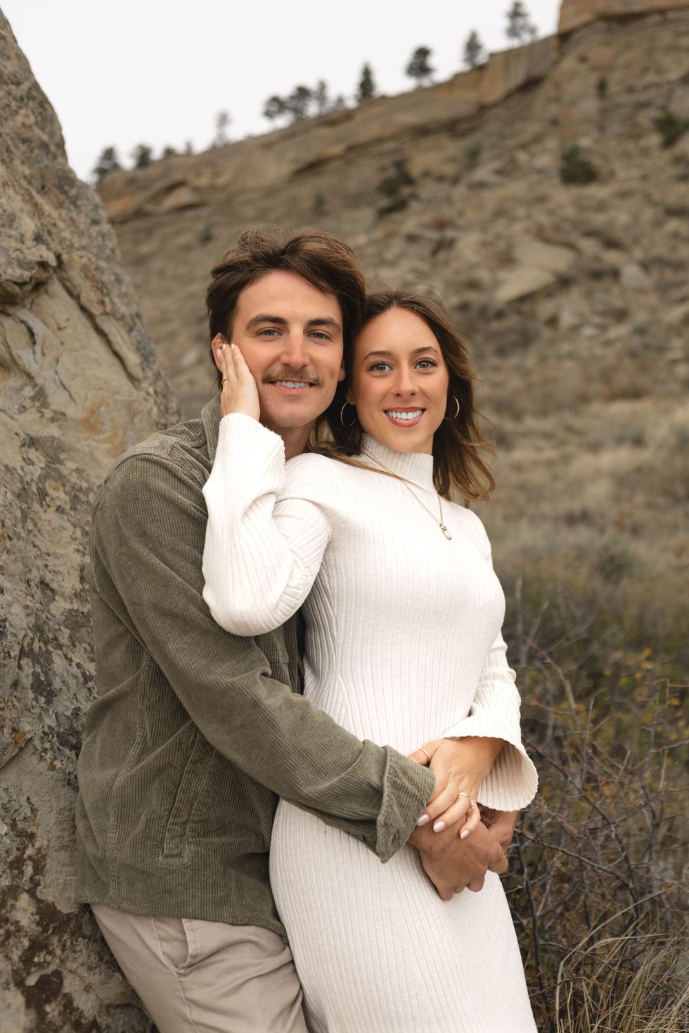 A couple embraces outdoors against a rocky hillside backdrop wearing white and olive colored clothing.