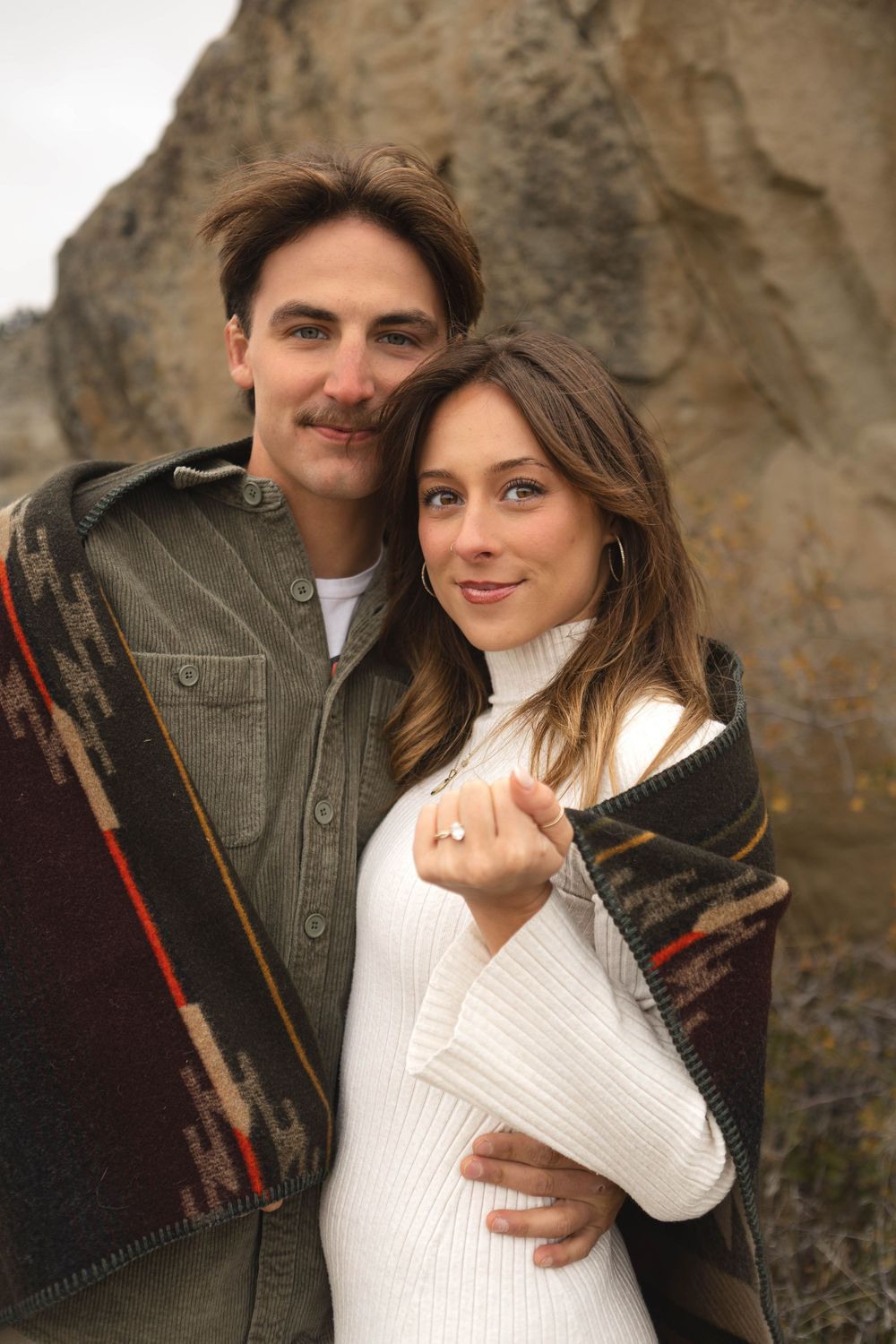 Romantic couple embraces outdoors against rocky backdrop wearing white sweater and dark jacket.