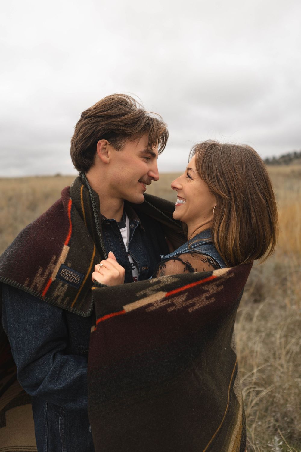 A couple sharing intimate moments in a field while wrapped in a plaid blanket on an overcast day.
