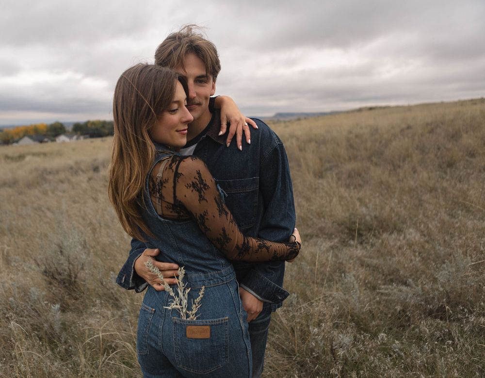 A couple embraces in denim clothing on a windswept hillside with moody overcast skies in the background.