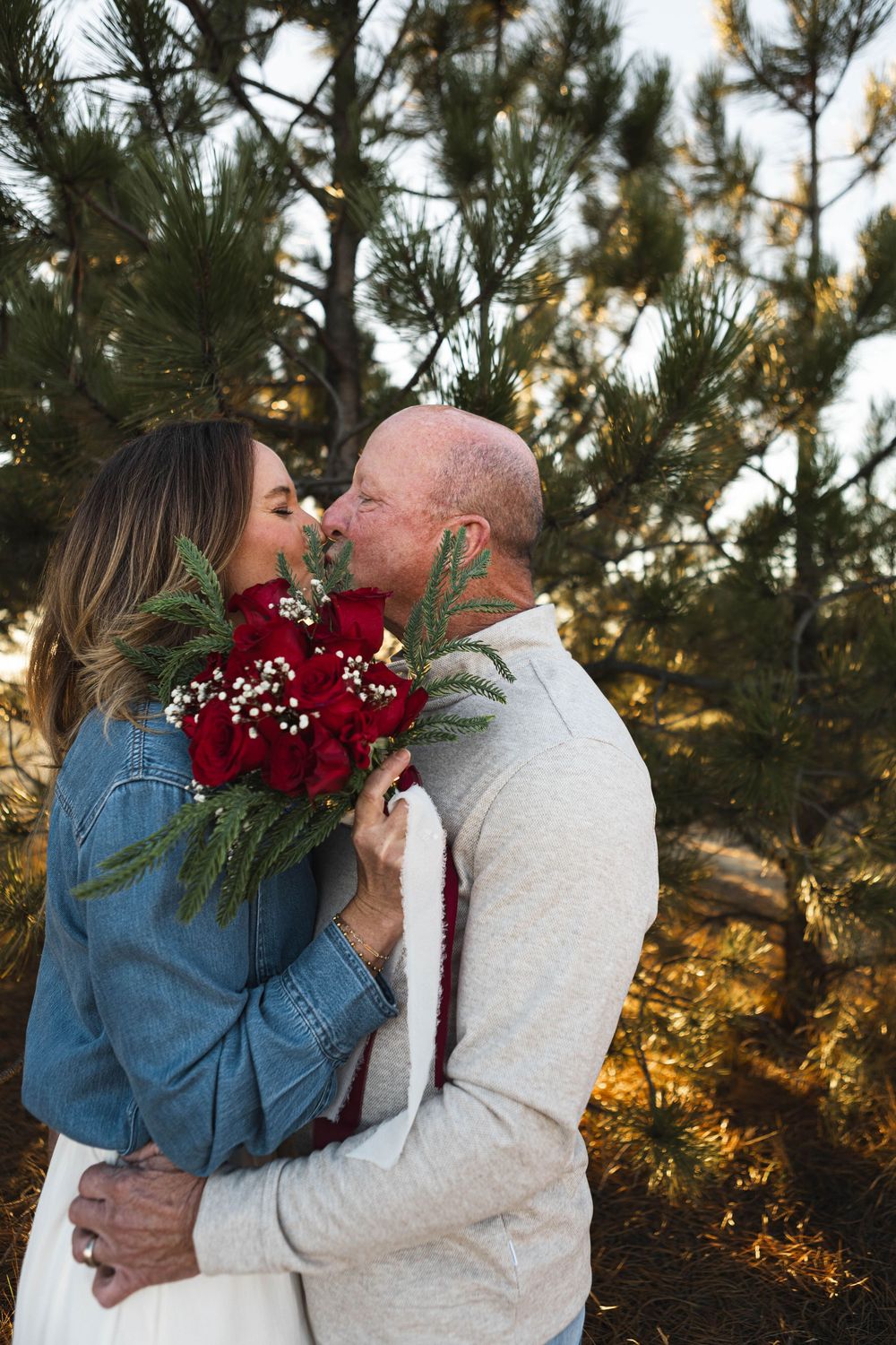 A couple embraces in a romantic outdoor setting with pine trees and golden sunlight while wearing denim and white.