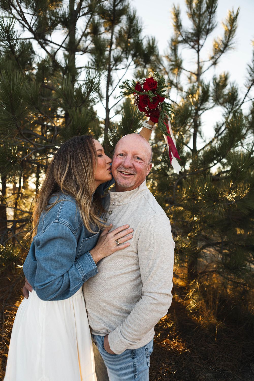 A couple shares a tender moment together in front of evergreen trees at sunset.