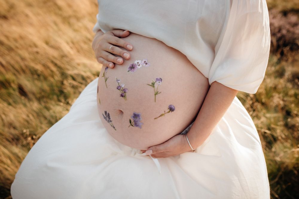 A pregnant person in a white dress cradles their belly while standing in a golden field at sunset.
