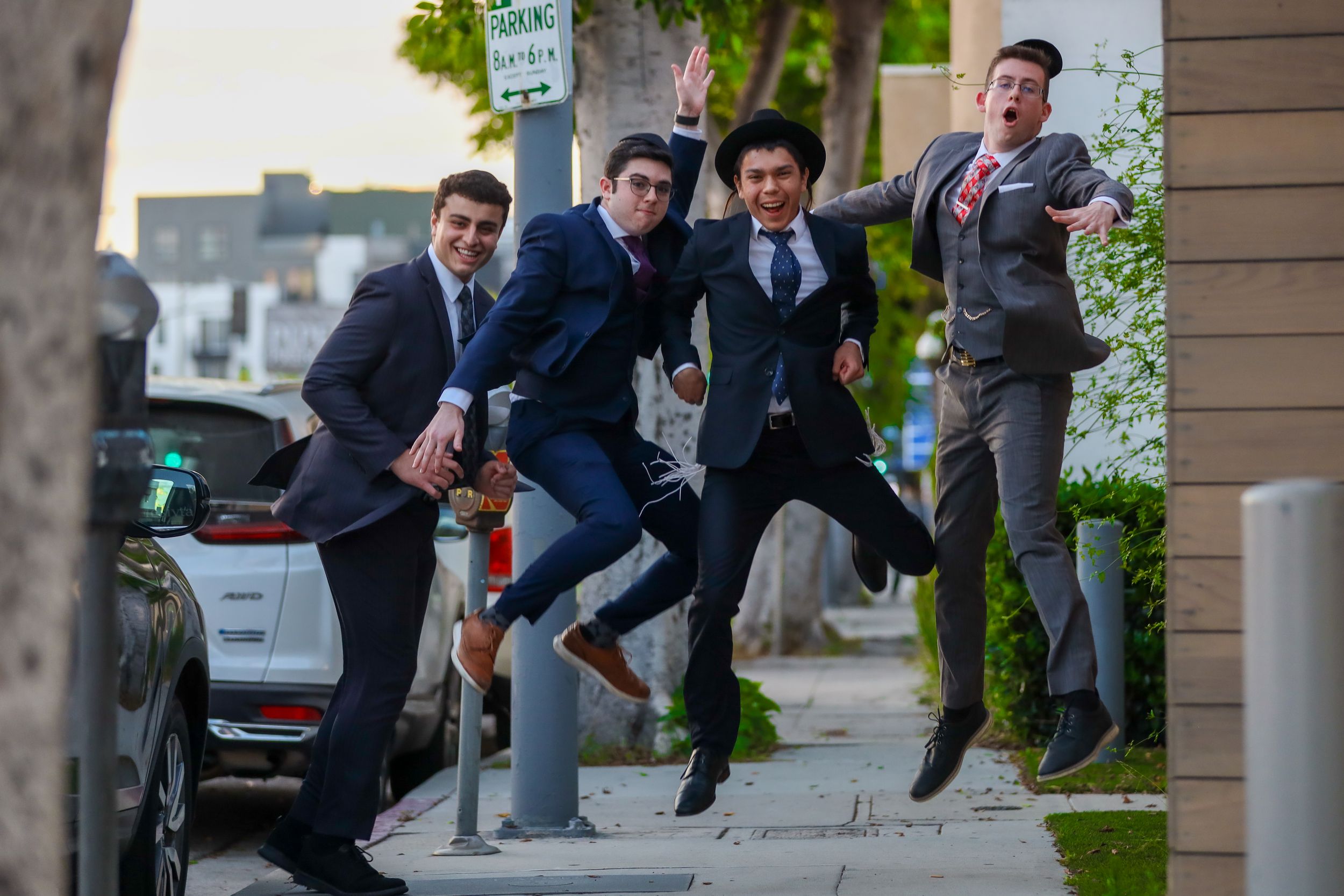 Four young men in suits jumping joyfully on a sidewalk in a candid outdoor photo.