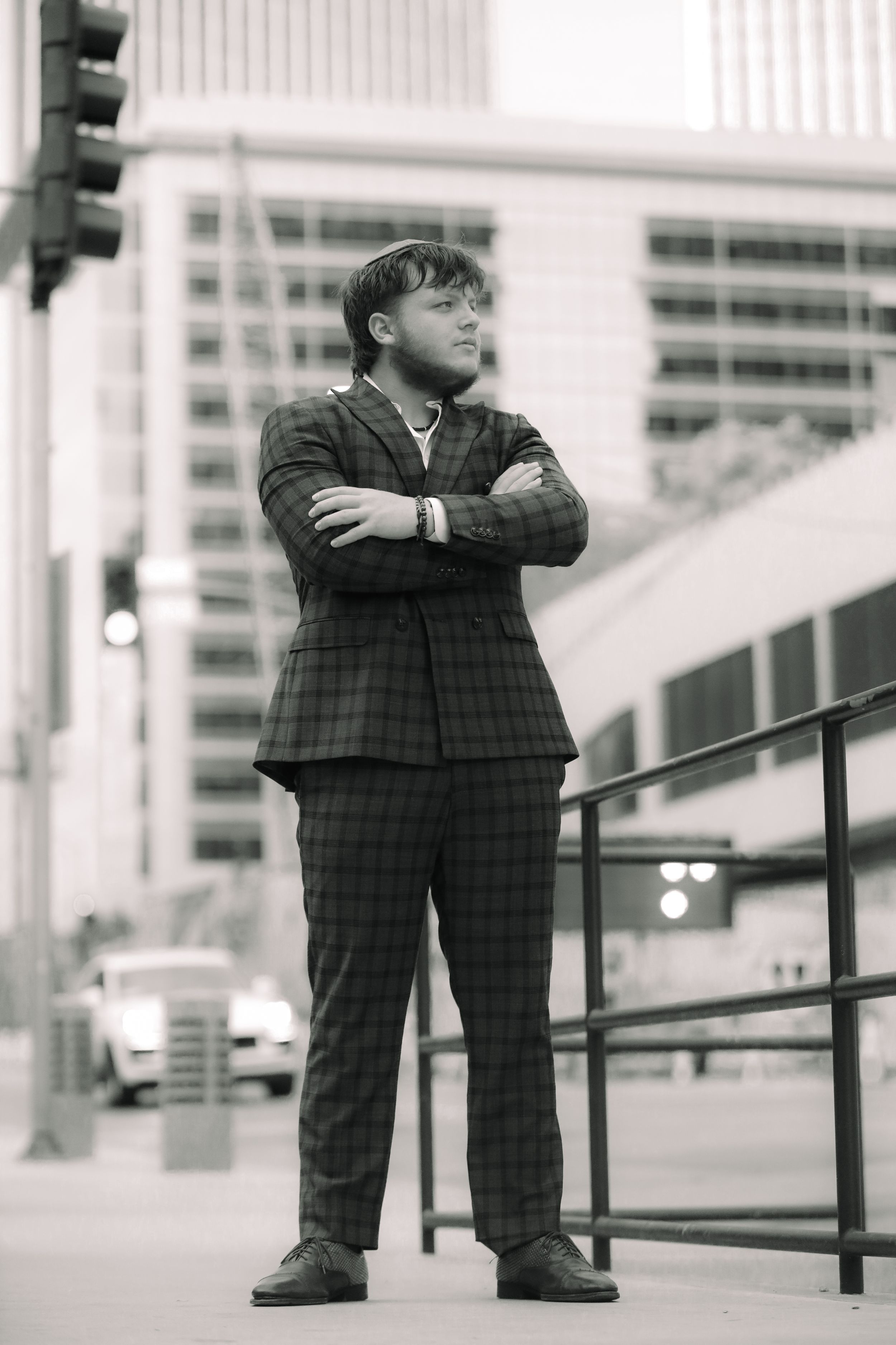 Young man in plaid suit stands with arms crossed on urban street in black and white.