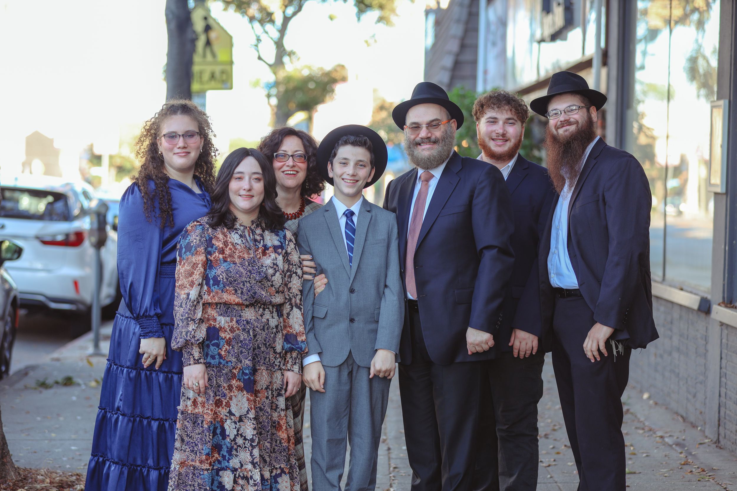 Family of seven smiles together on a sidewalk in semi-formal attire outdoors.