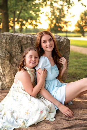 Two people wearing white and blue dresses pose together on a stone wall during golden hour in a park setting.