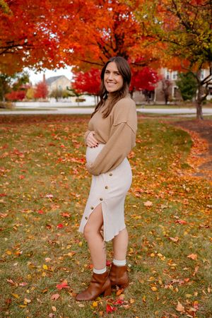 A woman poses playfully in a beige sweater and brown boots among vibrant red and orange fall foliage on a grassy lawn.