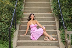 Someone in a flowing pink sundress sits casually on outdoor concrete stairs surrounded by greenery on a sunny day.