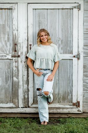 Young woman in mint top and ripped jeans posing against rustic white barn doors outdoors.