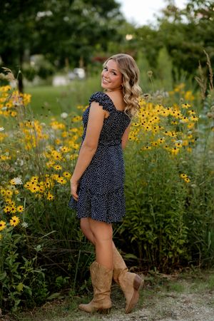 Young woman in navy polka dot dress and cowboy boots posing among yellow wildflowers in a lush garden.