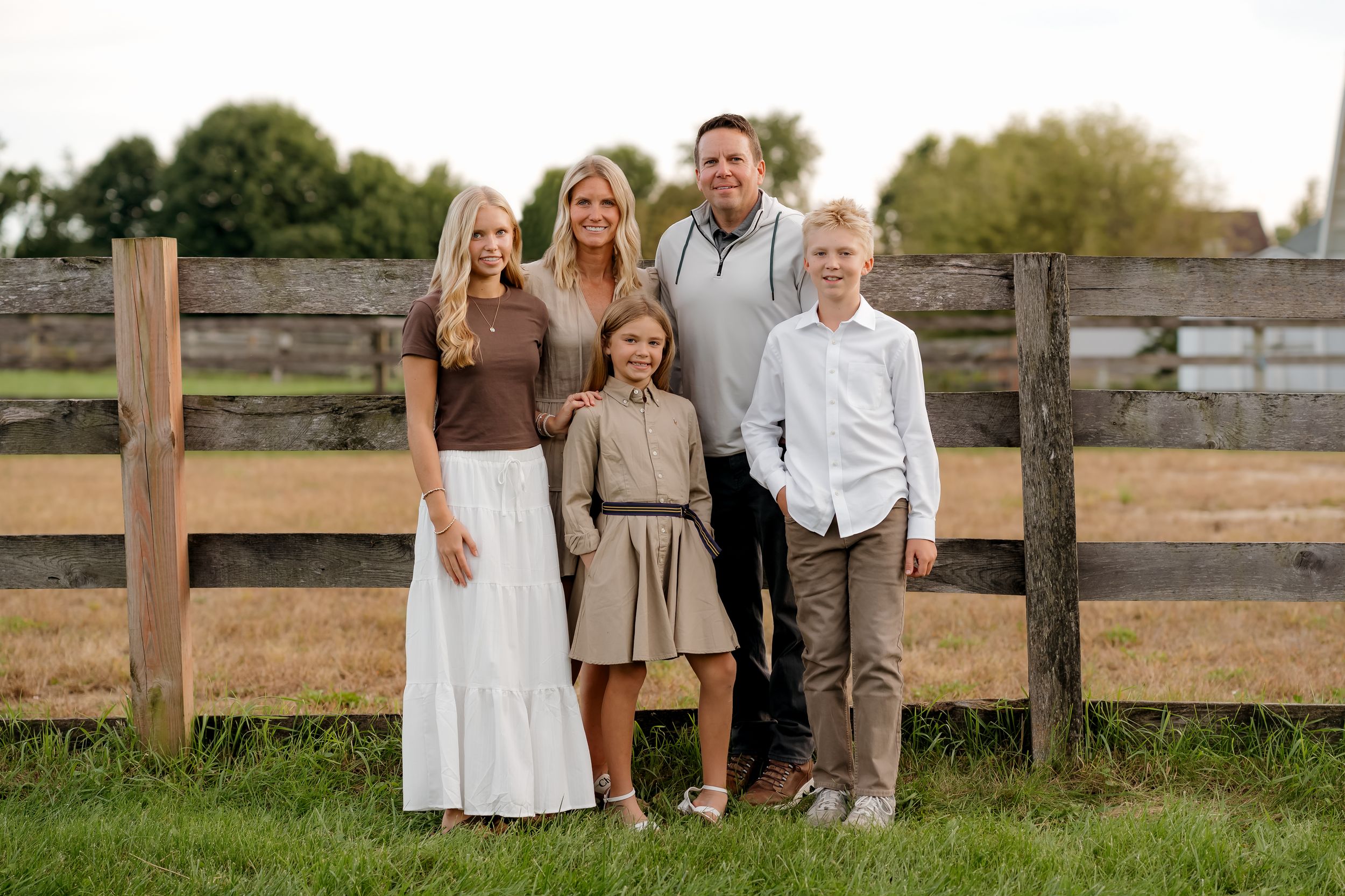 Family of five smiling together in front of a wooden fence on a green farm field during golden hour.