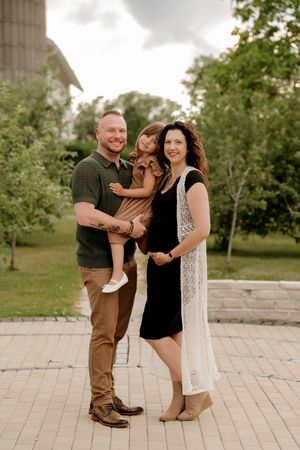 A smiling family of three poses outdoors, with a man holding a child and a pregnant woman beside them.