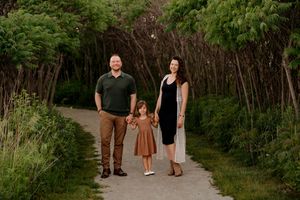 Family of three holding hands on a scenic tree-lined path, parents in casual attire with young daughter in brown dress.