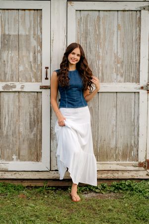 Young woman in denim top and white maxi skirt smiling against rustic weathered white barn doors.