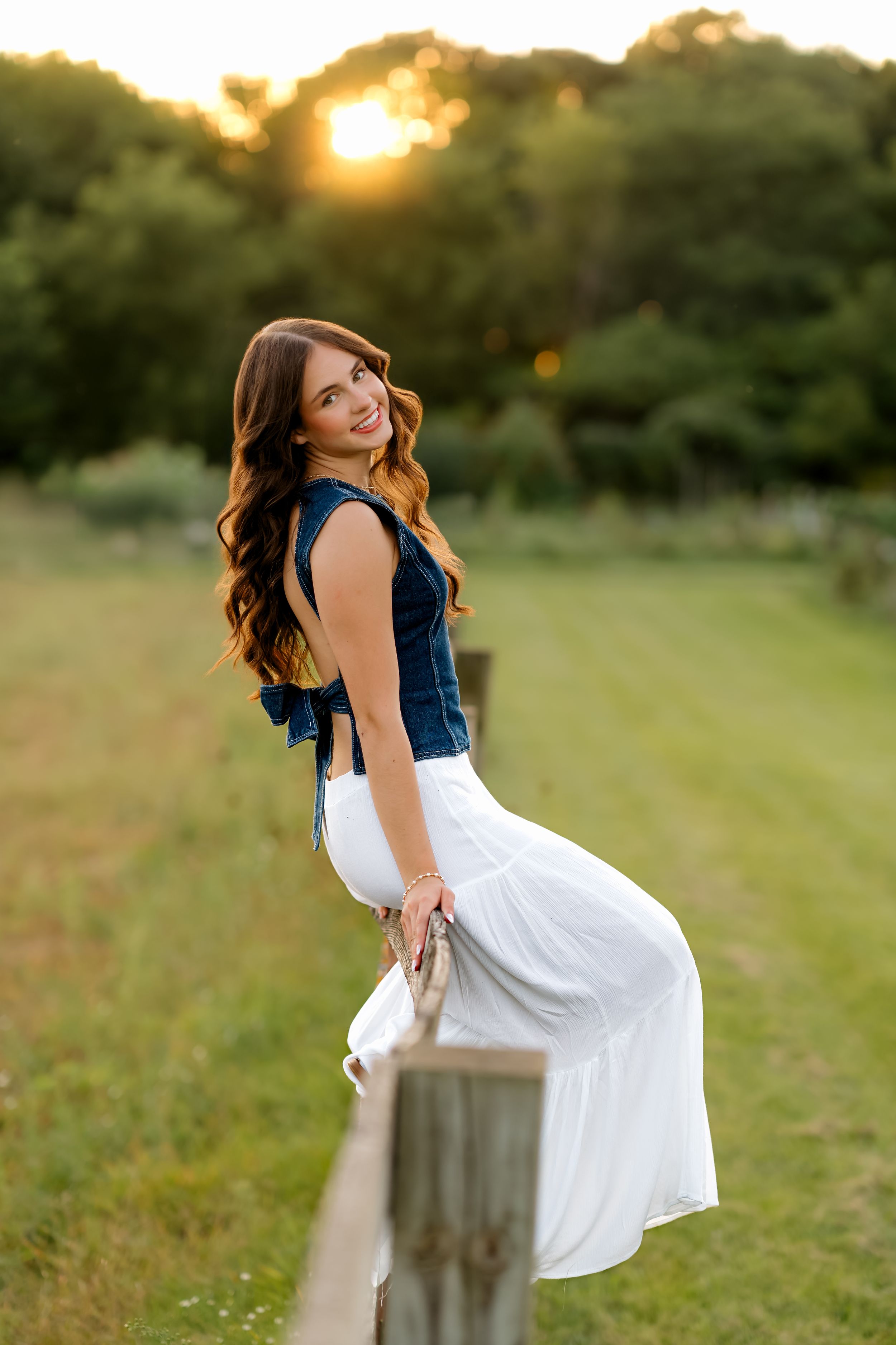 Young woman in denim top and white skirt smiling at sunset by a wooden fence in a lush green field.
