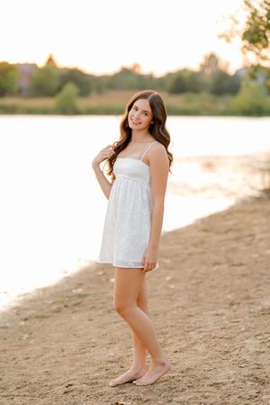 Young woman in white sundress standing barefoot on sandy beach shore at golden sunset.