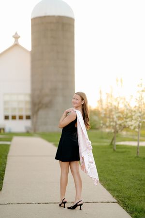 Young woman in black dress and heels holds white scarf on sidewalk near barn silo at golden hour.