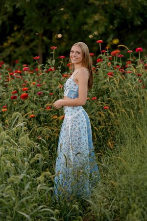 Young woman in blue floral maxi dress smiling in a lush garden with vibrant red flowers and green foliage.