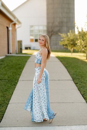 Young blonde woman in blue floral two-piece maxi skirt set posing on a sidewalk near a barn.