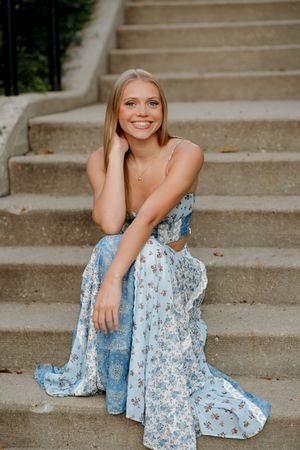 Young woman in blue floral maxi dress sitting on stone steps, smiling brightly outdoors.