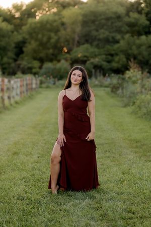 Young woman in elegant burgundy spaghetti-strap gown with slit poses gracefully in lush green garden pathway.