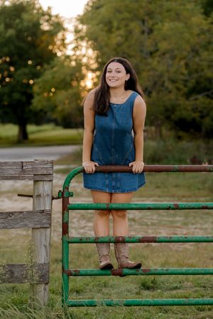 Young woman in denim dress and cowboy boots leaning on rustic green farm gate at golden hour.