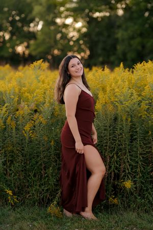 A smiling woman in a burgundy slit maxi dress poses barefoot in front of vibrant yellow wildflowers at golden hour.