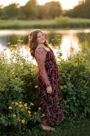 Young woman in floral burgundy gown stands among wildflowers by a reflective pond at golden hour sunset.