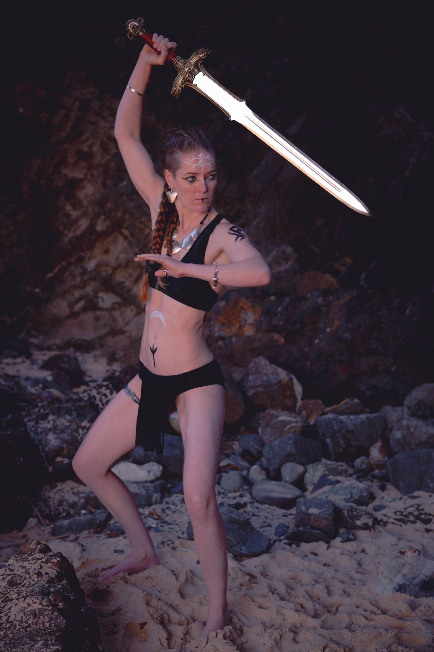 Viking figure in black athletic wear poses with raised sword on beach at night.