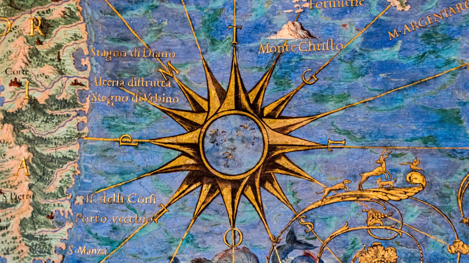 Artistic macro photography of an antique nautical map featuring a large golden compass rose and radiating rhumb lines over a blue ocean.