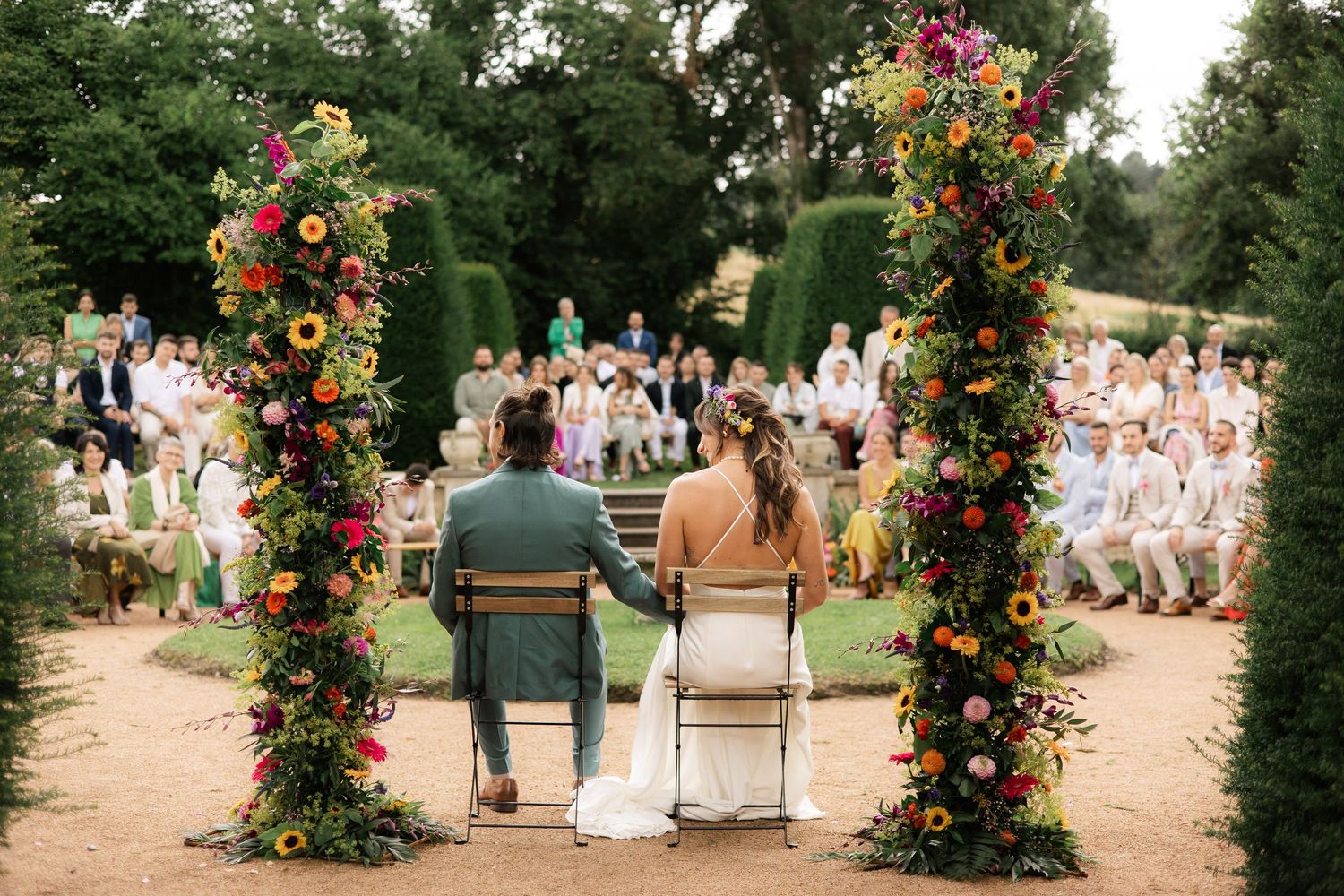 Photo d'un couple pendant leur cérémonie laïque. Ils sont face à leurs invités entouré d'une arche fleurie.