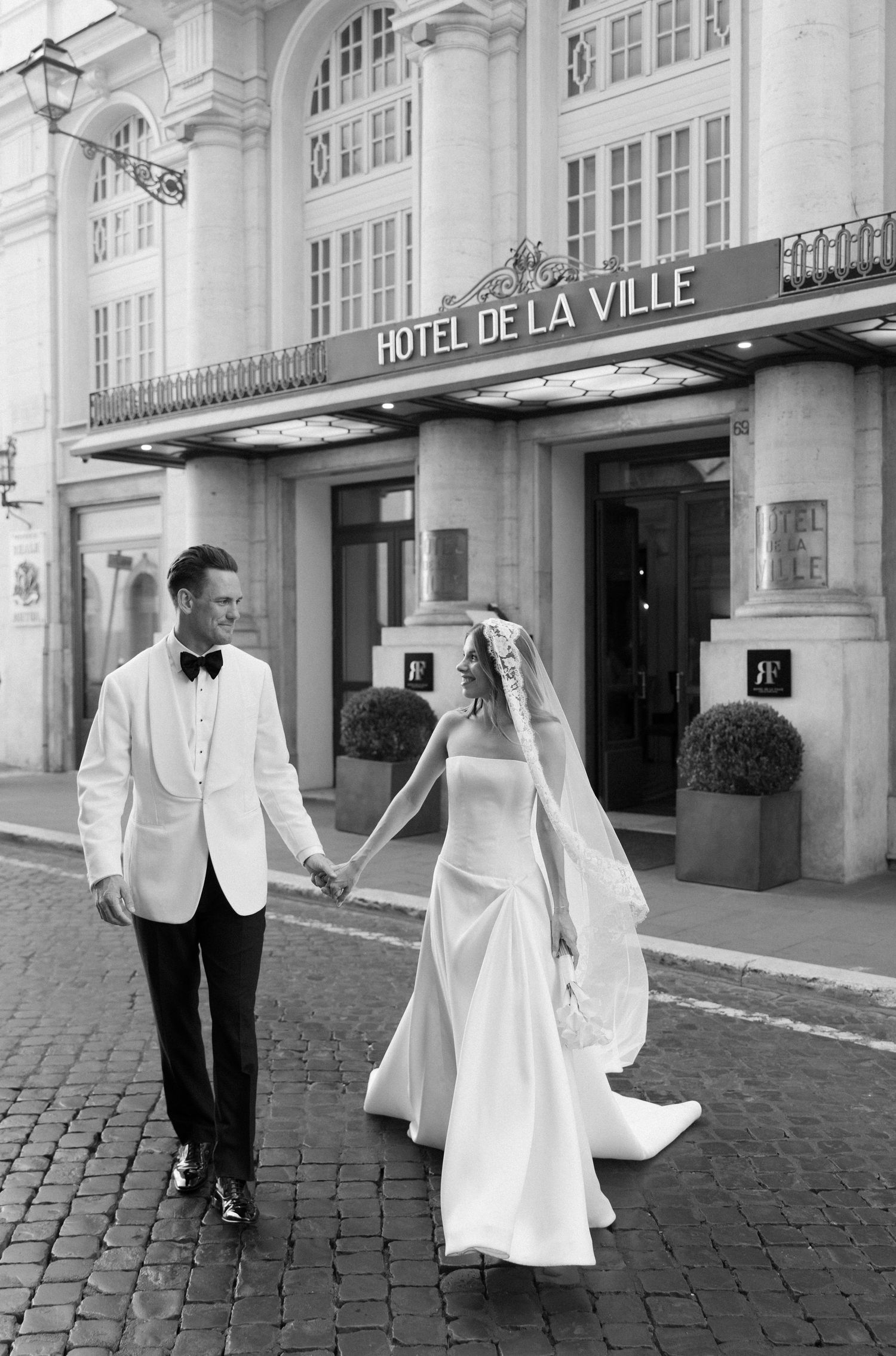 A bride and groom walk together outside Hotel de la Ville, with classic architecture and cobblestone streets in view.