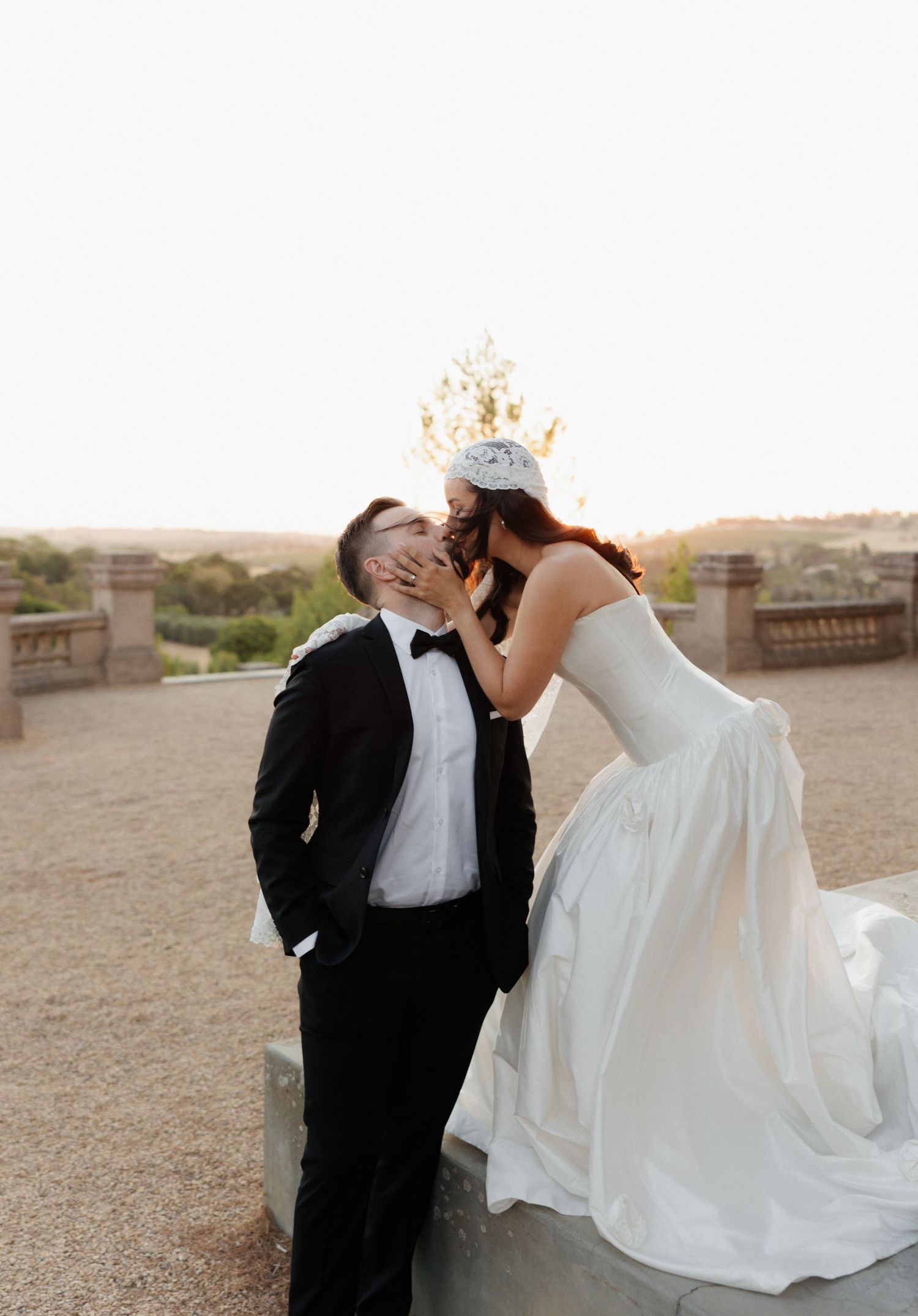 A romantic wedding couple shares an intimate moment in their formal attire against a desert landscape at sunset.