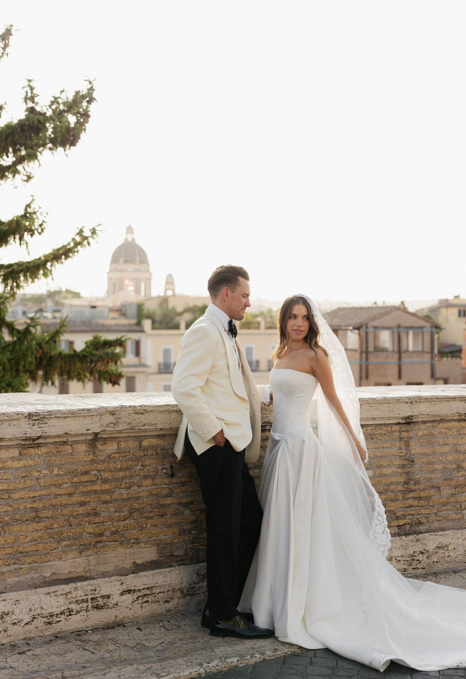 Romantic couple in wedding attire embracing on a stone wall overlooking historic Roman architecture at sunset.
