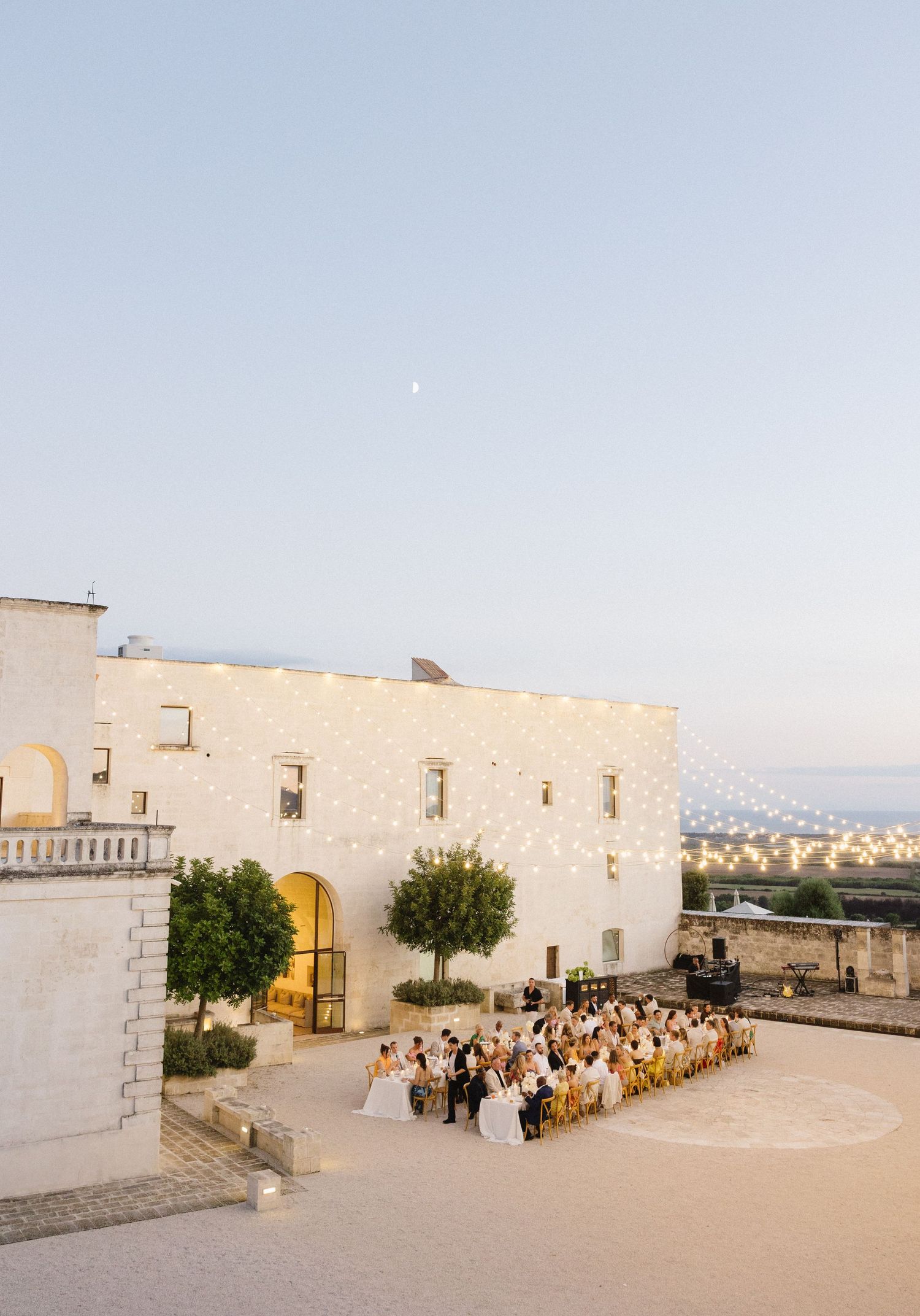 Group of people gathered in the courtyard of a white Mediterranean building during a wedding ceremony at sunset.