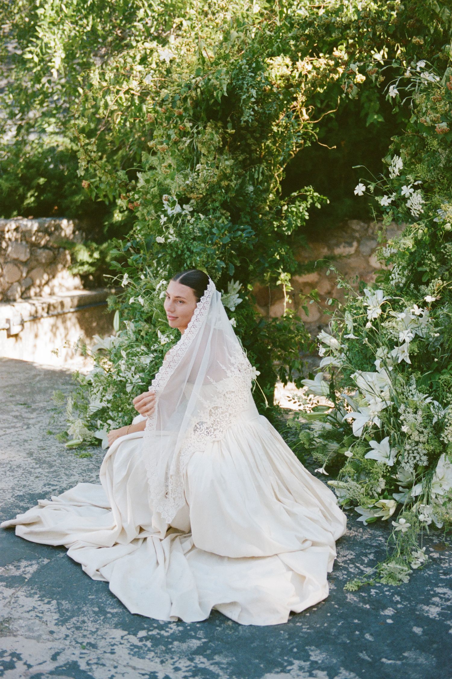 Bride in a white gown and veil sits gracefully among lush green floral arch outdoors on her wedding day.