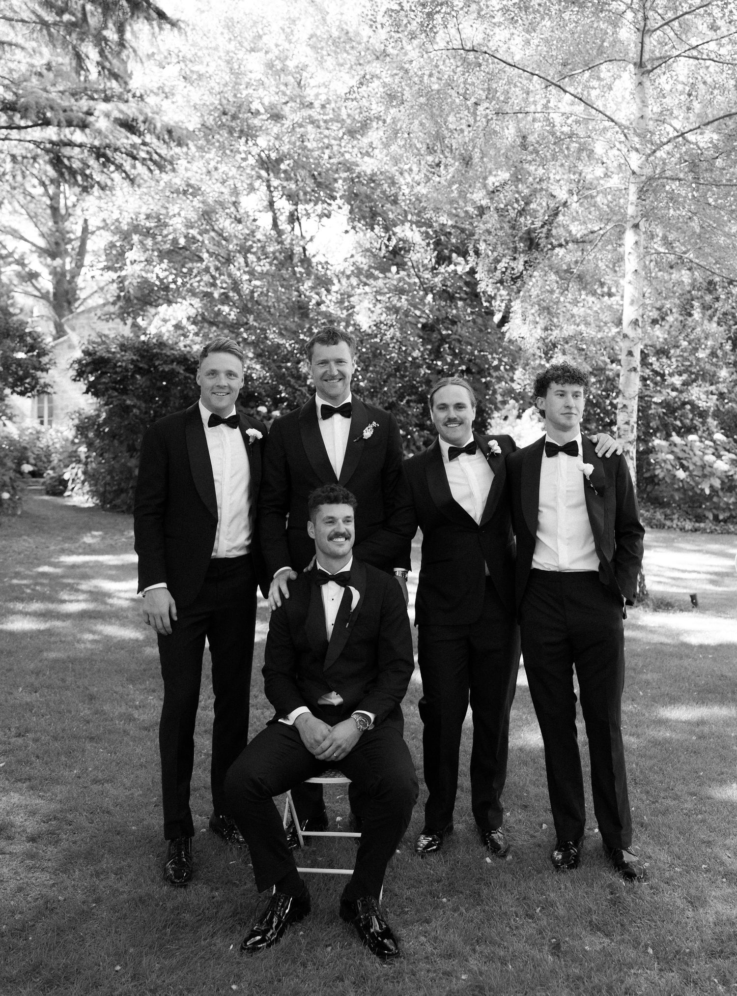 A group of groomsmen in black tuxedos pose together outdoors for wedding photos in a natural setting.