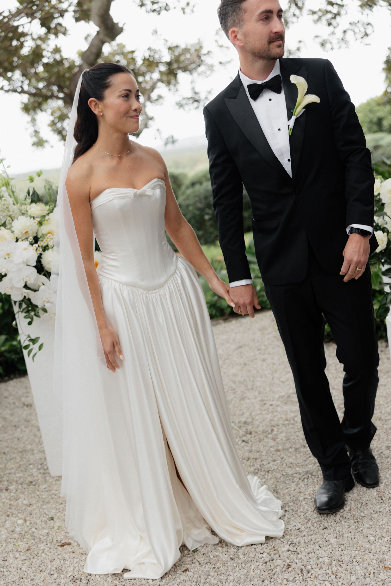 Bride in a strapless white satin gown with a veil holds hands with a groom in a black tuxedo and bow tie on a gravel path.
