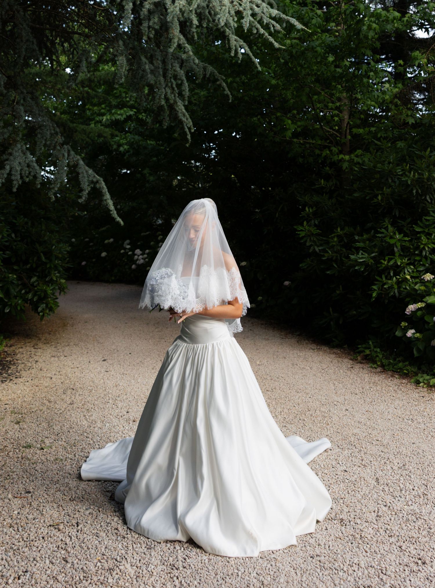A bride in a classic white wedding gown and veil stands on a gravel path surrounded by lush green trees.