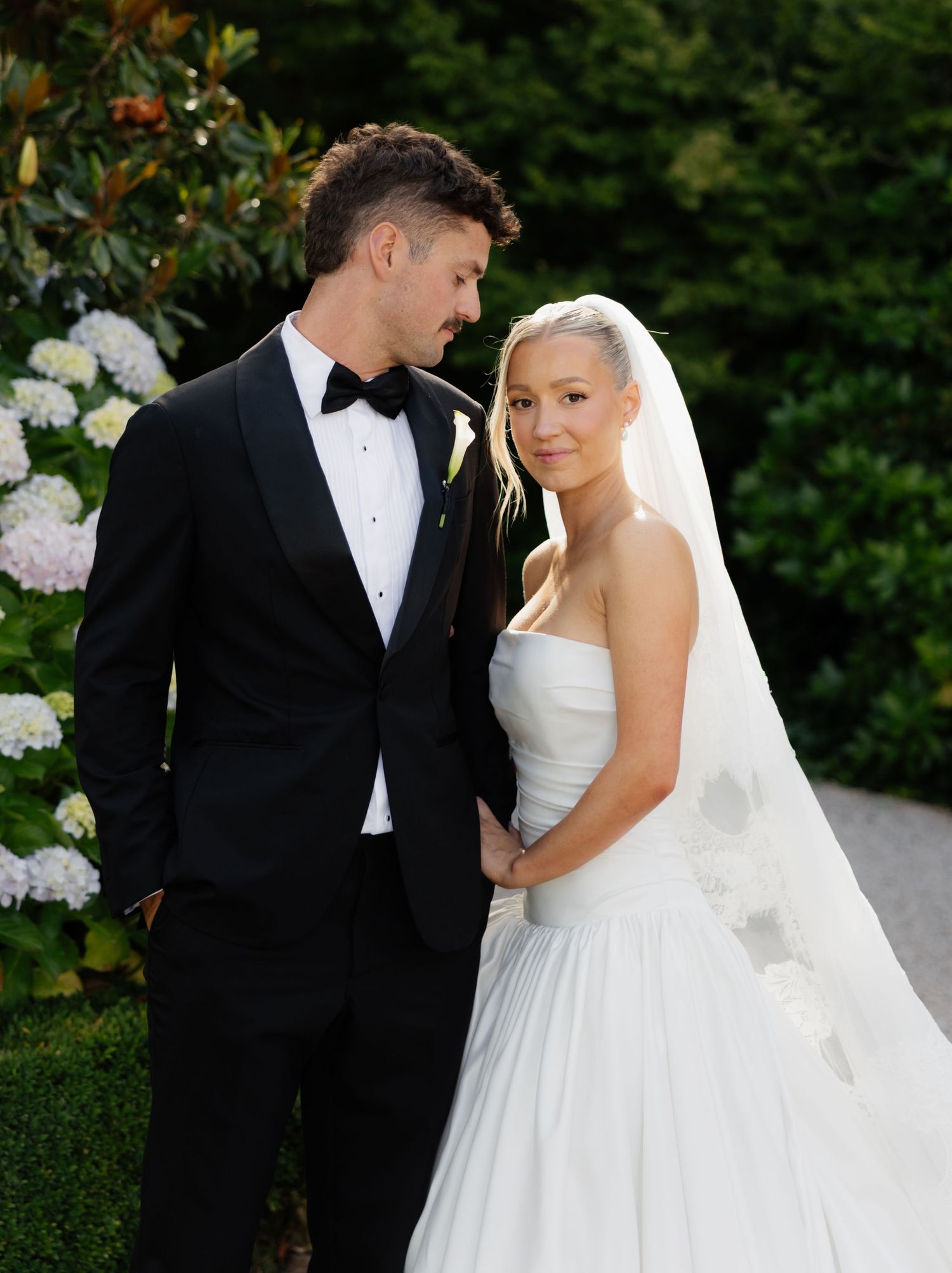 Romantic wedding couple shares intimate moments in a garden setting wearing classic black tuxedo and white bridal gown.