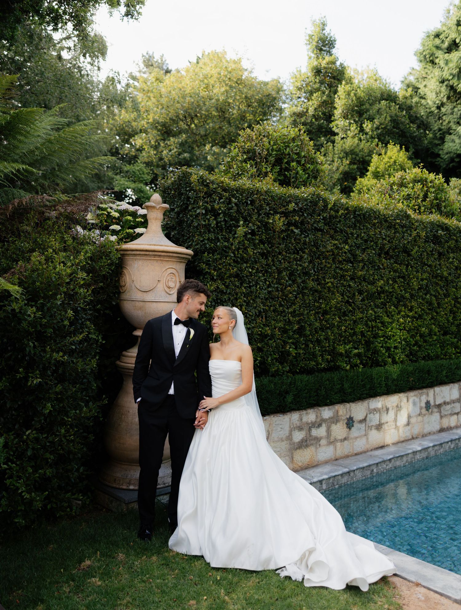 A couple in formal wedding attire shares an intimate moment by a swimming pool surrounded by manicured hedges.