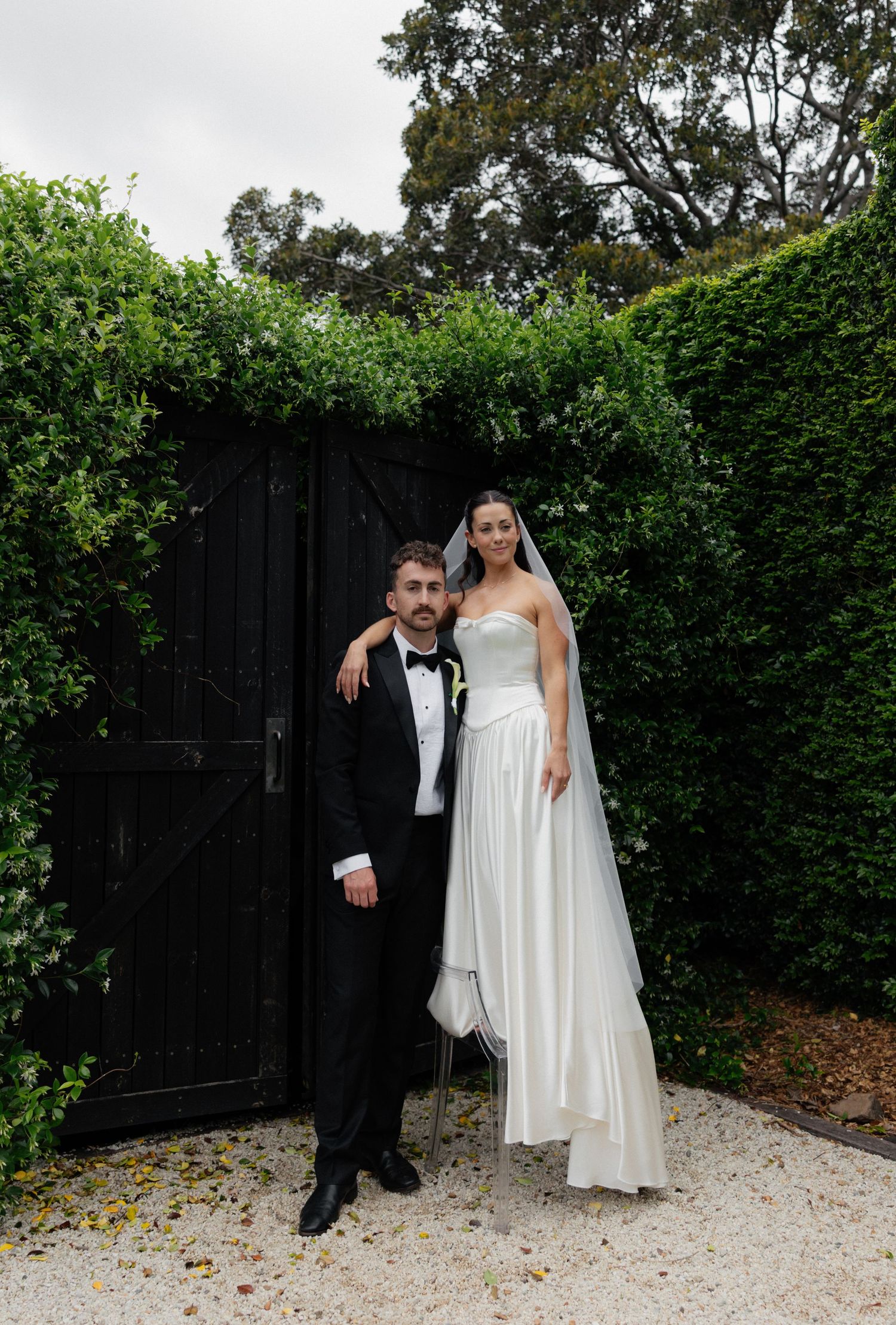 Bride in a strapless white gown and veil stands beside a groom in a black tuxedo and bow tie before a black gate and green hedge.