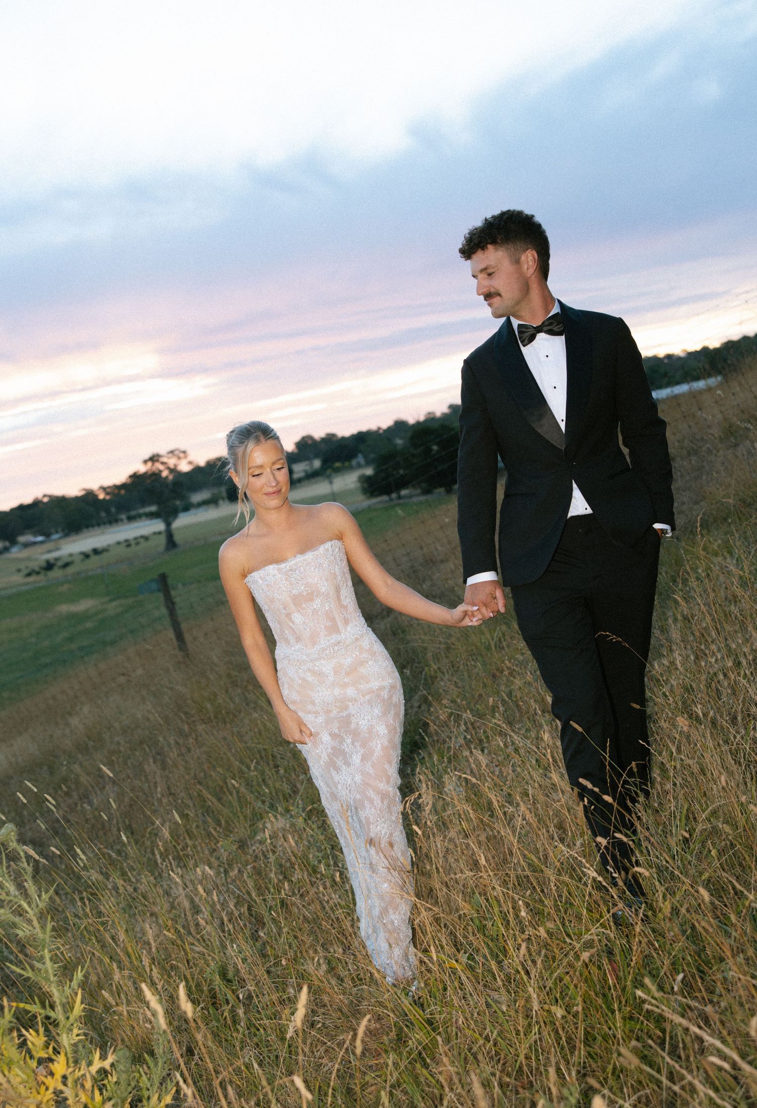 A couple in wedding attire poses together in a field of tall grass during sunset.