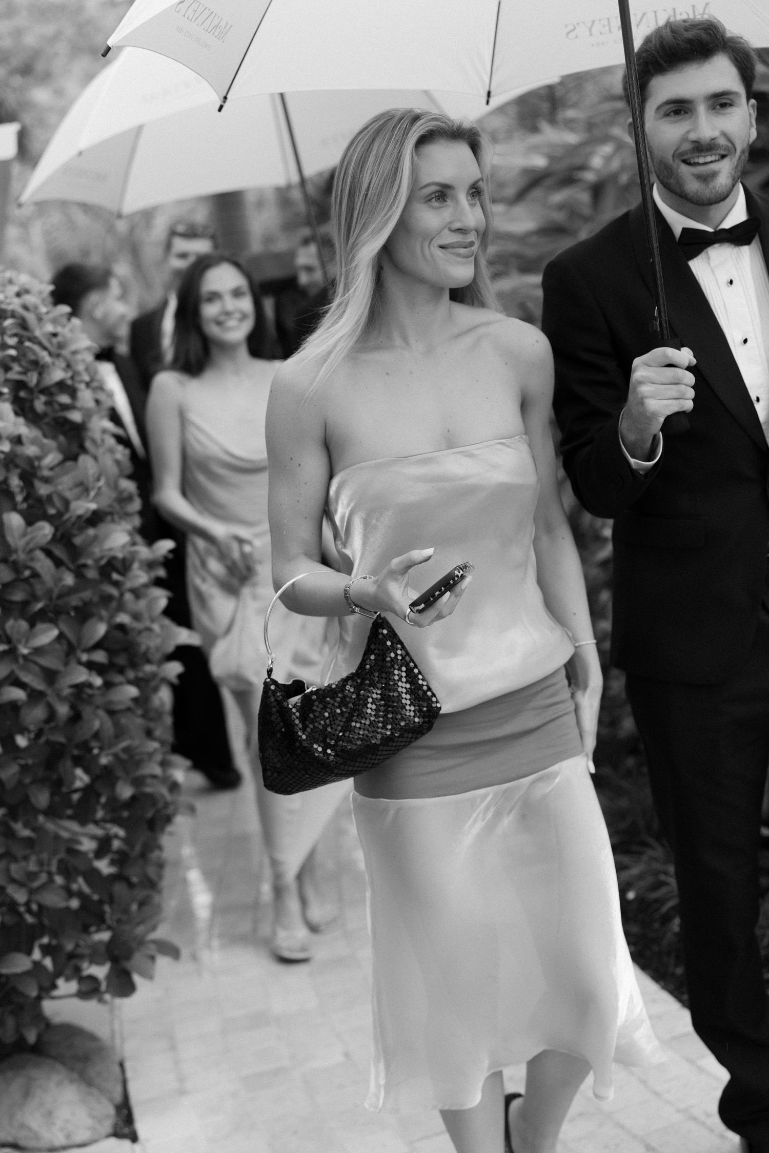 Black and white photo of wedding guests in formal attire walking under white umbrellas along a garden path, a woman holding a sparkly clutch