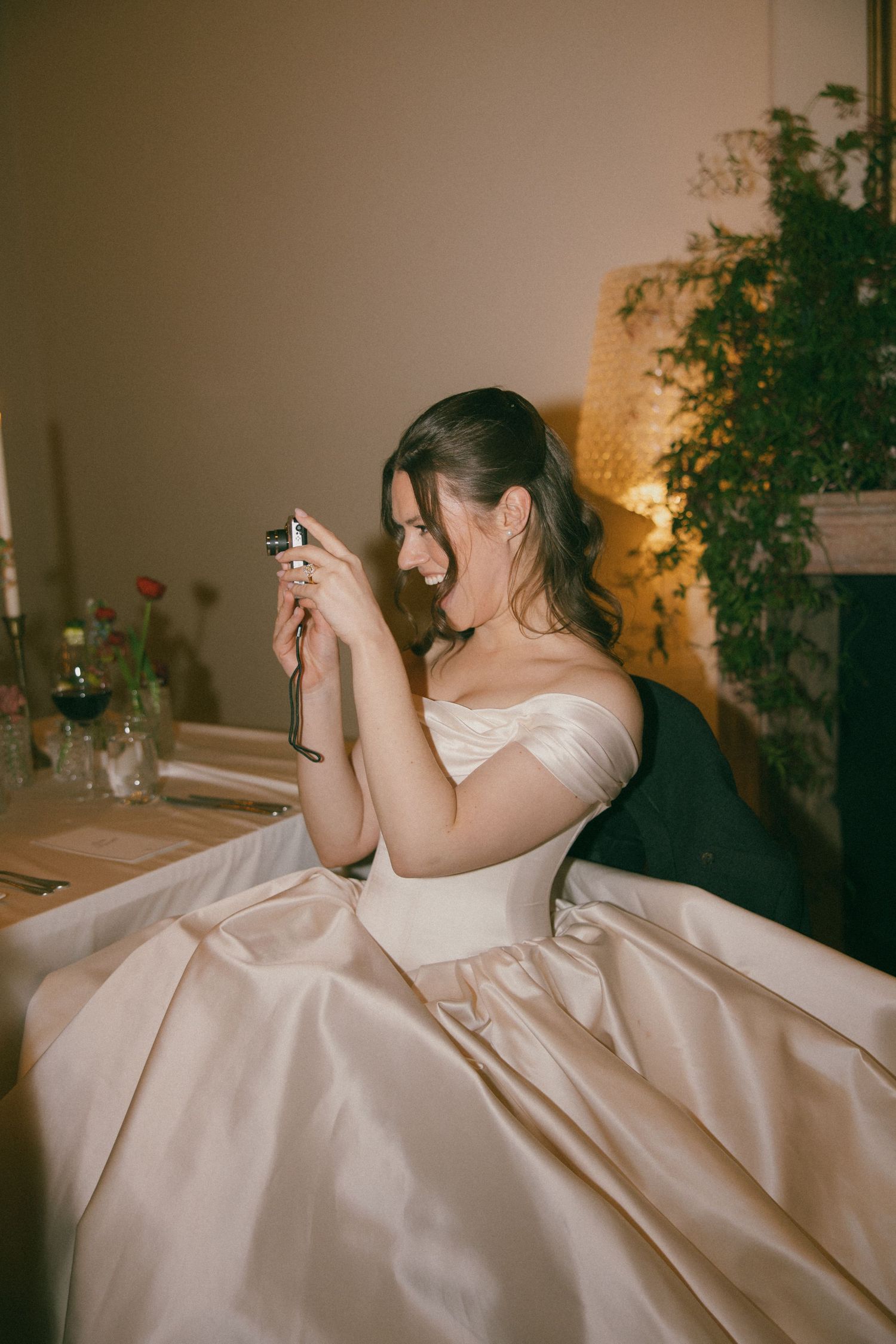 Bride in off-shoulder satin ballgown sits at reception table, smiling while taking photos with a small camera.