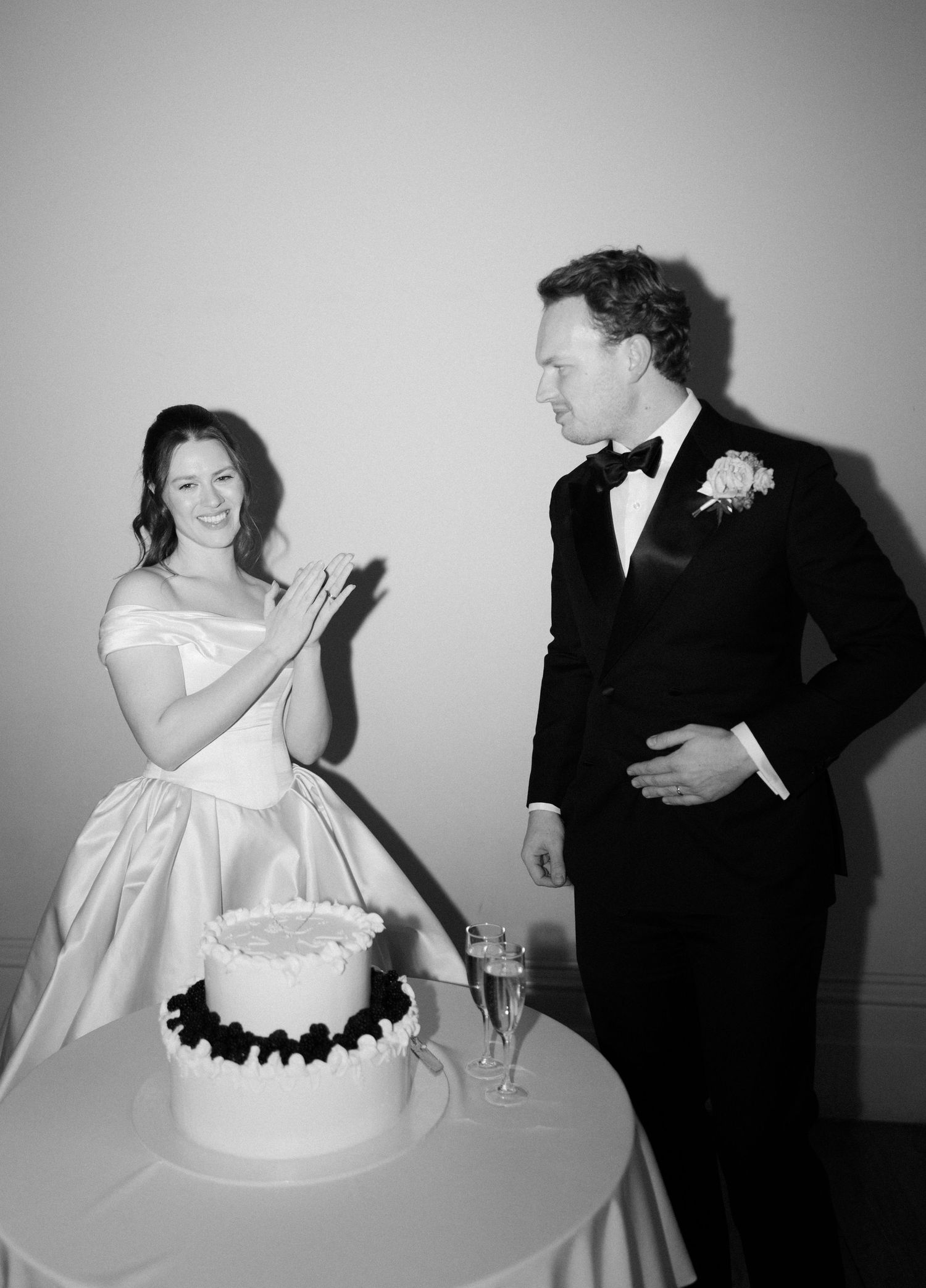 Black and white photo of bride and groom smiling beside wedding cake at elegant reception.